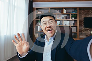 Funny Asian businessman looks at the camera of a smartphone, talks on a video call, a man works in the office, waves