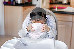 Funny Asian Chinese kid girl sitting in high chair eating drinking soup from plate bowl. Healthy eating for kids children. Toddler