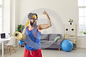 Funny athlete holding gym mat and dumbbells and showing his weak biceps after sport workout at home