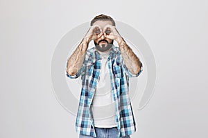 Funny attractive bearded man showing binocular with hand gesture, looking very concentrated, over gray background. Happy