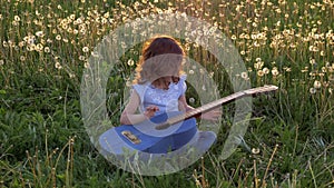 Funny attractive little girl sits in the grass and plays the jeans guitar. field with dandelions in spring bloom at sunset. concep