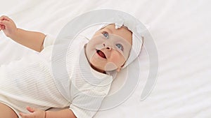 a funny baby with blue eyes of six months lies on the back and smiles on the bed in a white bodysuit in a bright bedroom