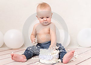 Funny baby boy eating cake witn balloons on the background. Dirty kid face and hands in the cream. first birthday celebration