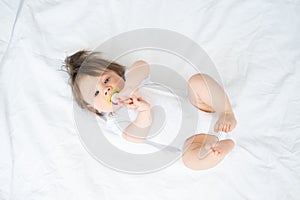 funny baby boy with nipple in bodysuit smiling and lying on a white bedding at home. top view.