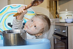 Funny baby child getting messy eating cereals or porridge by itself with a wooden spoon, straight from the cooking pot