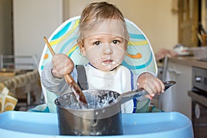 Funny baby child getting messy eating cereals or porridge by itself with a wooden spoon, straight from the cooking pot