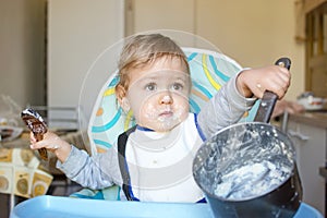Funny baby child getting messy eating cereals or porridge by itself with a wooden spoon, straight from the cooking pot