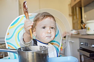 Funny baby child getting messy eating cereals or porridge by itself with a wooden spoon, straight from the cooking pot