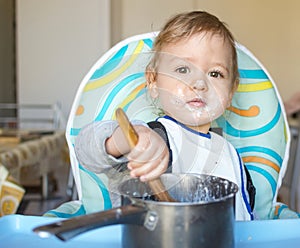 Funny baby child getting messy eating cereals or porridge by itself with a wooden spoon, straight from the cooking pot