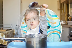 Funny baby child getting messy eating cereals or porridge by itself with a wooden spoon, straight from the cooking pot
