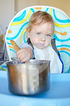 Funny baby child getting messy eating cereals or porridge by itself with a wooden spoon, straight from the cooking pot