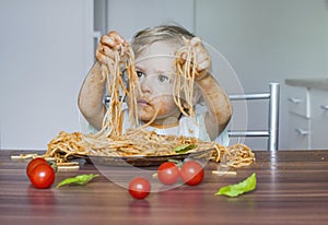 Funny baby child getting messy eating spaghetti with tomato sauce from a large plate, by itself with his hands, at home