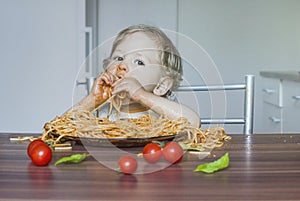 Funny baby child getting messy eating spaghetti with tomato sauce from a large plate, by itself with his hands, at home