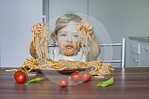 Funny baby child getting messy eating spaghetti with tomato sauce from a large plate, by itself with his hands, at home