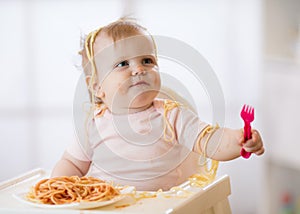 Funny baby eating noodle. Grimy kid girl eats spaghetti with fork sitting on table at home photo