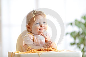 Funny baby eating noodle. Grimy kid eats spaghetti with handssitting on table at home