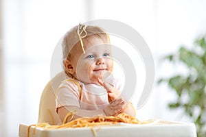Funny baby eating noodle. Grimy kid eats spaghetti with handssitting on table at home photo
