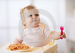 Funny baby eating noodle. Grimy kid girl eats spaghetti with fork sitting on table at home