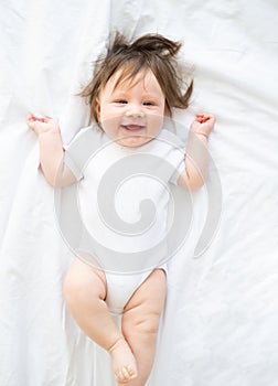 funny baby girl in white bodysuit smiling and lying on a white bedding at home. top view.