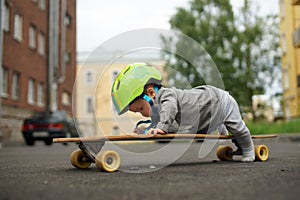 Funny baby in green helmet lying on belly on skateboard and pushing himself by one leg. Little skateboarder