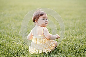 Funny baby kid. Cute adorable baby girl in yellow dress sitting on grass in park outdoor and looking back. Funny child toddler