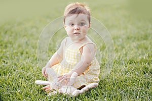 Funny baby kid with toy. Cute adorable baby girl in yellow dress sitting on grass in park outdoors. Funny child toddler playing