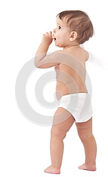 Funny baby stays near wall and holds finger in a mouth.