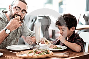 Funny beaming son feeling happy eating cheesy pizza with father