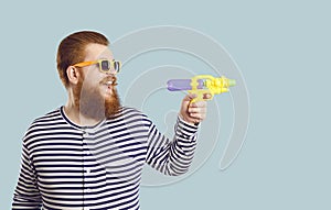 Funny bearded chubby male tourist shoots with small water pistol towards copy space.