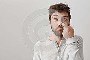 Funny bearded guy with messy hair and bad habits, staring at camera with stupid face, picking in nose with index finger