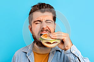 Funny bearded man enjoying cheat meal savoring burger, blue background