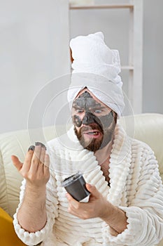 Funny bearded man having fun with a cosmetic mask on his face made from black clay. Men skin care, humor and spa at home