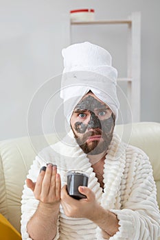 Funny bearded man having fun with a cosmetic mask on his face made from black clay. Men skin care, humor and spa at home