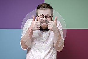 Funny bearded man in round glasses, makes an approving gesture with hands, thumbs up, closes eyes and grins teeth.