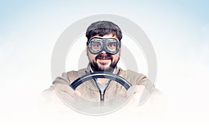 Funny bearded man in stylish goggles with steering wheel, smoke around, light blue background. Front view. Car driver concept