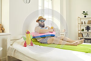 Funny bearded man with travel accessories lying in bed imagining resort vacation