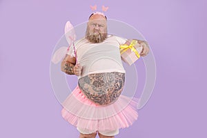 Funny bearded plump man in fairy costume with gift box grimaces on purple background