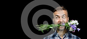 Funny bearded stylish man holding bouquet of wild flowers in his mouth on black background. Greeting card, gift for your loved one