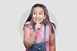 Funny beautiful brunette young girl in casual pink t-shirt, blue overalls standing, telling secret, toothy smile and looking away