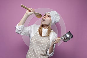 Funny beautiful girl cook in apron holds dishes and sings on a pink background, crazy woman housewife holds kitchen items and