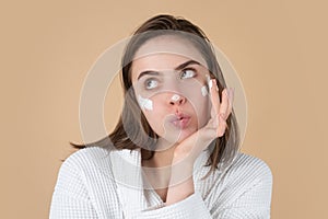 Funny beautiful woman applying moisturizer cream on her pretty face. Female skin care, close up face beauty portrait.