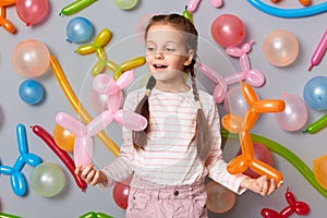 Funny birthday kid. Playing time. little girl with braids standing against gray wall decorated with colorful balloons, smiling