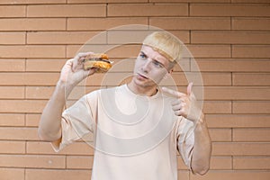 A funny blond European man dressed in a beige T-shirt holding a burger