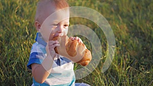 Funny blonde toddler boy eating tasty bread sitting in green grass outdoor.