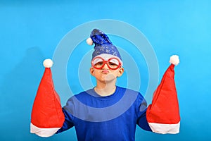 A funny boy in a blue Santa Claus hat holds red hats for New Year and Christmas in his hands