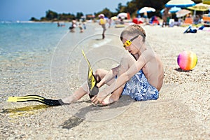 Funny boy diver sitting on sandy beach putting on diver flippers