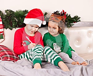 Funny boy kid in Santa hat and girl in deer mask writing letter to Santa Claus sitting on a bed