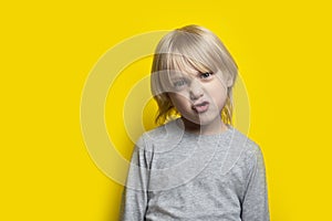 Funny boy with long blond hair poses faces. Portrait of child on yellow background