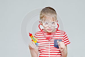 Funny boy playing doctor. Toy syringe, glasses and phonendoscope. Portrait