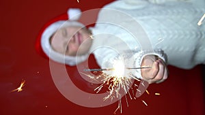 funny boy in Santa Claus cap holding sparkler on the red background